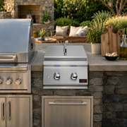 A stainless steel outdoor power burner with a lid and two control knobs mounted on a stone-tiled outdoor kitchen counter, with a matching grill to its left and kitchen decor in the background.