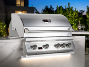 A stainless steel outdoor gas grill installed on a marble-topped patio counter, with control knobs, a digital display, and an illuminated under-shelf light. The background shows a modern house, trees, and a twilight sky.