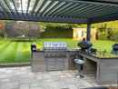 Outdoor kitchen island under a slatted patio cover, featuring a stainless steel gas grill, a black wood-fired pizza oven, gray stone counters, and a single bar stool, set on a stone patio with a green lawn in the background.